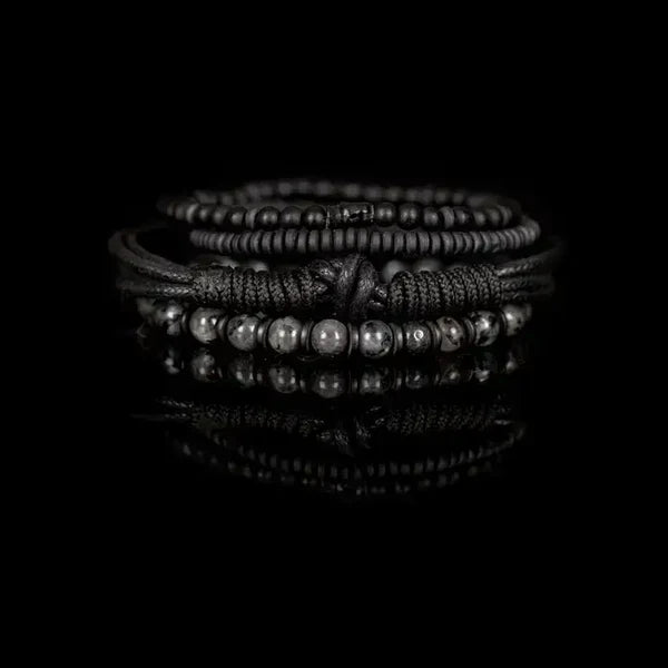 Men's layered black bracelet set featuring matte black beads, natural gray stone beads, braided leather cords, black wood beads, and hematite spacers in a rugged contemporary luxury design.