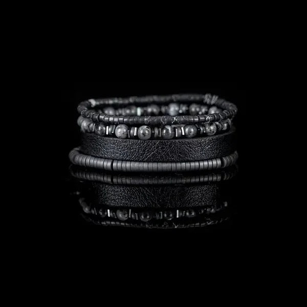 Layered men’s bracelet set featuring a black leather cuff, matte black hematite disc beads, gray natural stone beads, and metallic accent spacers, displayed on a white background.