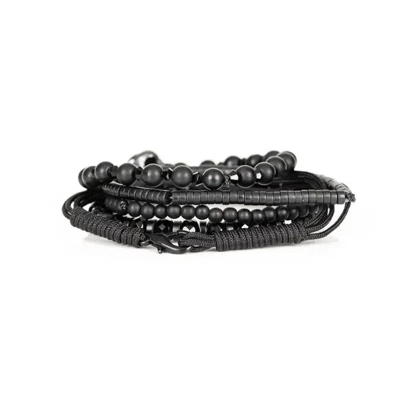 Black layered men’s bracelet with matte beads, faceted black stones, braided cord, and silver skull centerpiece on a white background.