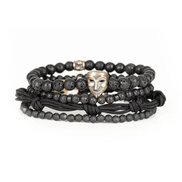 Black layered men’s bracelet featuring lava stone beads, faceted black stone accents, braided cord wraps, and an antique gold theatrical mask centerpiece on a white background.