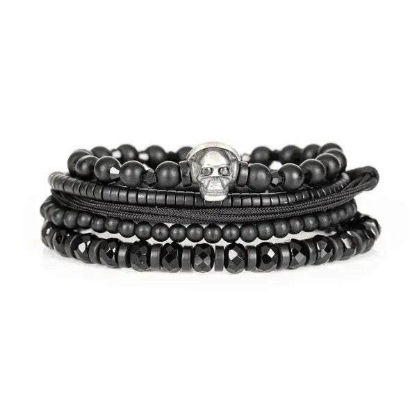 Black layered men’s bracelet with matte beads, faceted black stones, braided cord, and silver skull centerpiece on a white background.