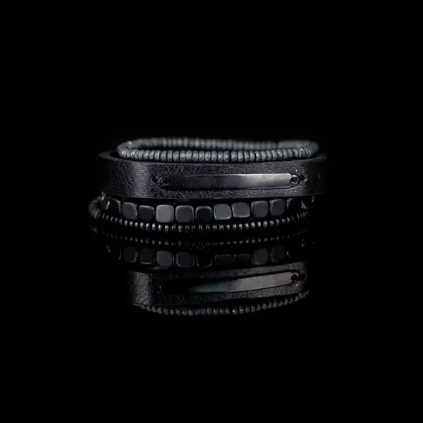Layered men’s bracelet set featuring a black leather band with a black metal plate, matte hematite cube beads, and hematite disc beads in a sleek monochromatic design, displayed on a white background.