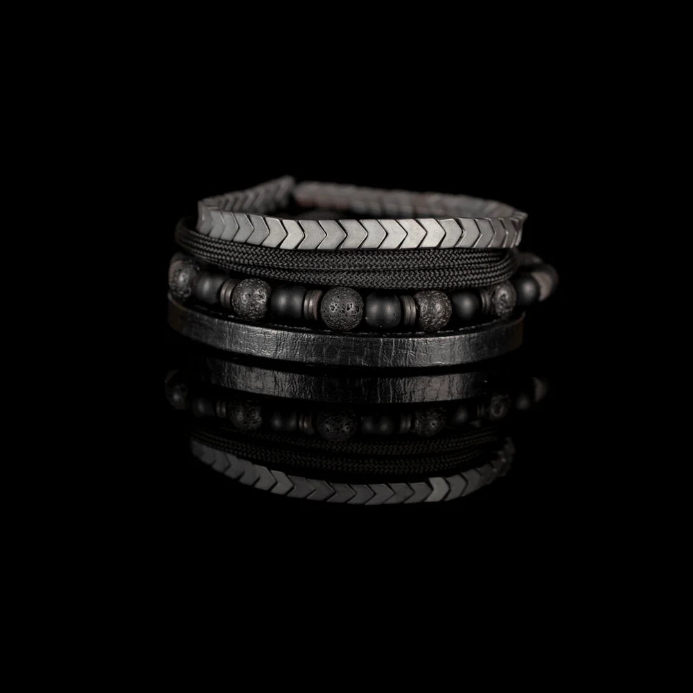 Men's handcrafted layered bracelet set featuring matte black onyx beads, black lava stone beads, black braided cord, genuine black leather band, and gunmetal hematite arrow-shaped beads. Premium minimalist men's bracelet stack photographed on a clean white background.