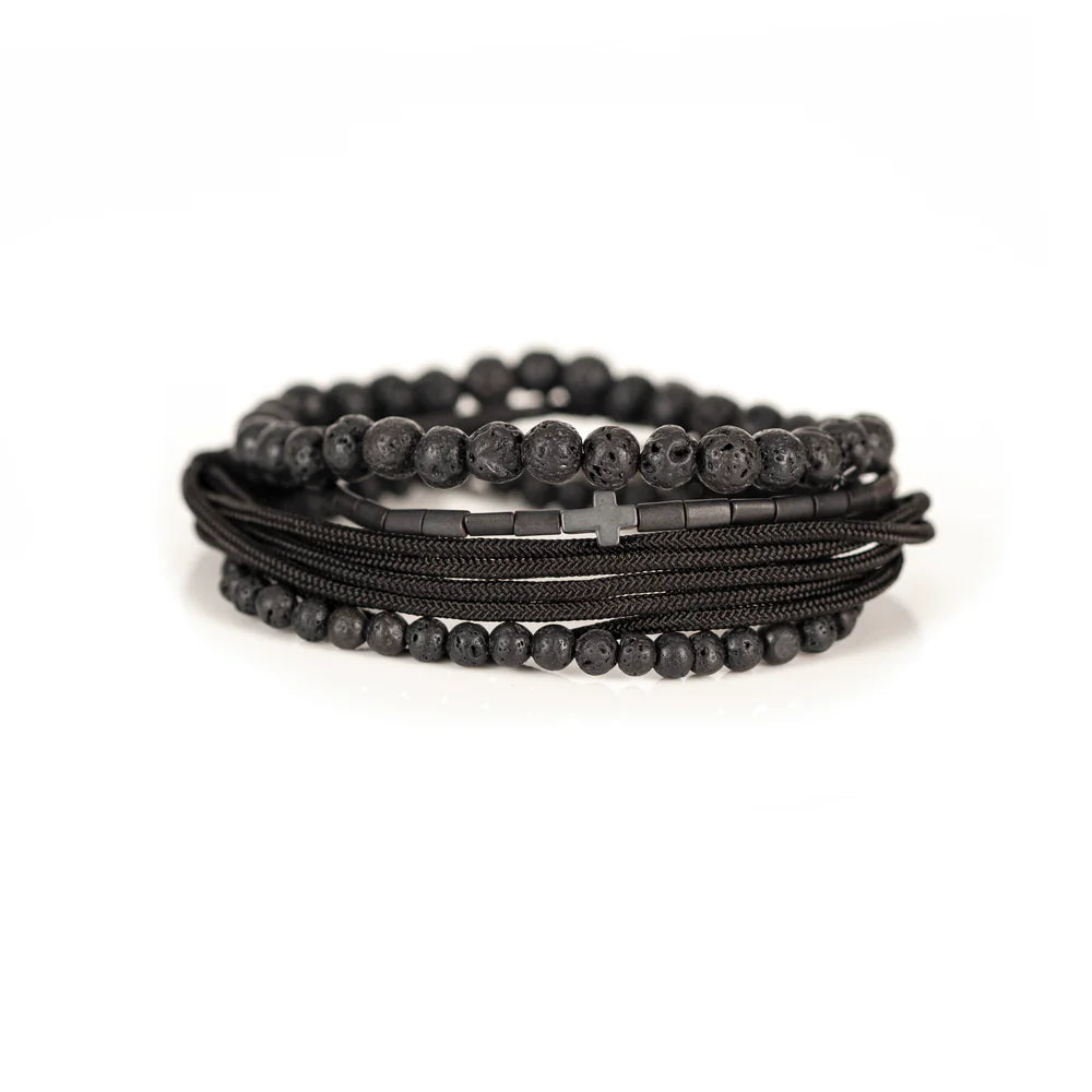 Men's handcrafted layered bracelet set featuring natural black lava stone beads, matte black hematite tube beads with a black cross accent, black braided cord, and black seed bead bracelets. Premium minimalist men's bracelet stack photographed on a clean white background.