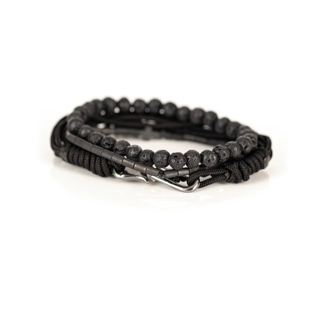 Men's handcrafted layered bracelet set featuring natural black lava stone beads, matte black hematite tube beads with a black cross accent, black braided cord, and black seed bead bracelets. Premium minimalist men's bracelet stack photographed on a clean white background.