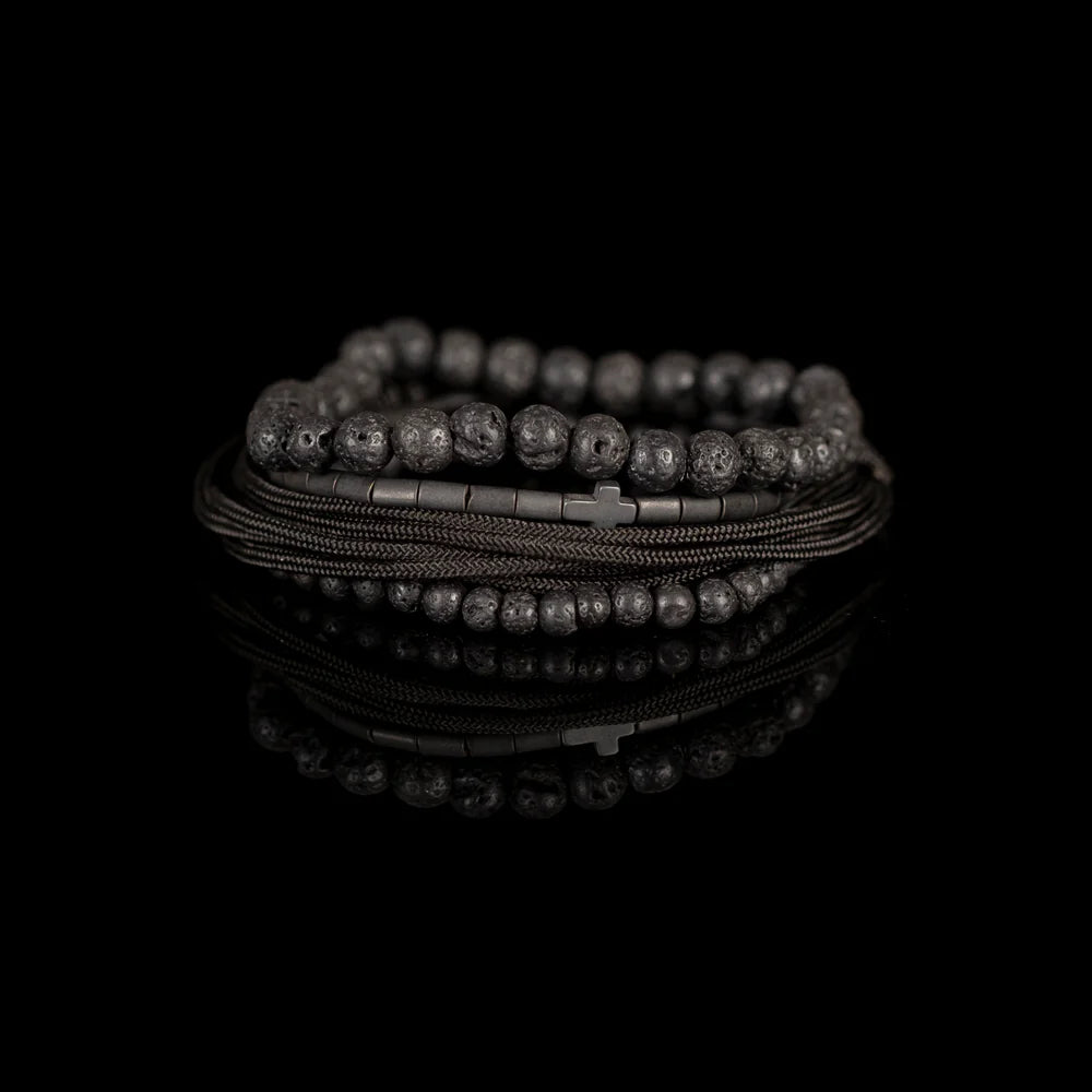 Men's handcrafted layered bracelet set featuring natural black lava stone beads, matte black hematite tube beads with a black cross accent, black braided cord, and black seed bead bracelets. Premium minimalist men's bracelet stack photographed on a clean white background.