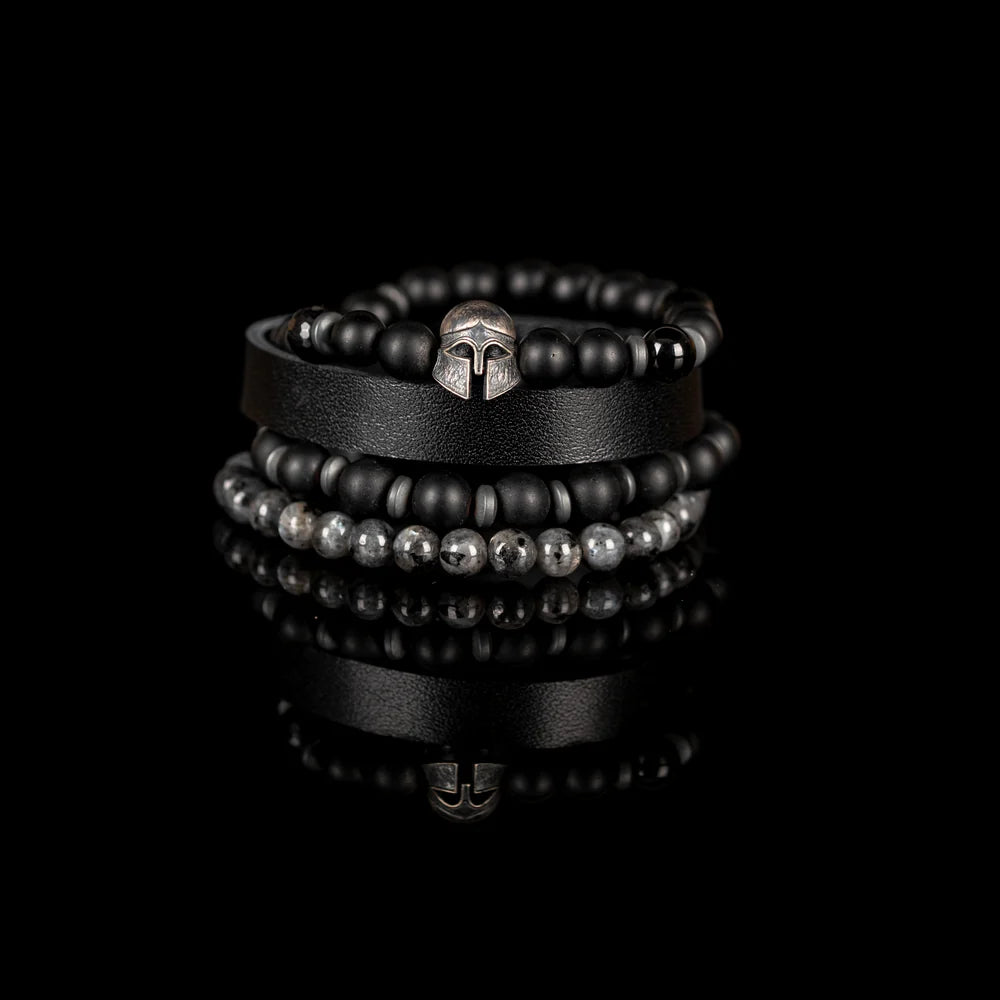 Men's handcrafted layered bracelet set featuring a black genuine leather cuff, matte black onyx beads, faceted black hematite beads, natural gray jasper gemstone beads, black hematite spacer beads, and a silver Spartan helmet centerpiece. Premium stacked men's bracelet photographed on a clean white background.