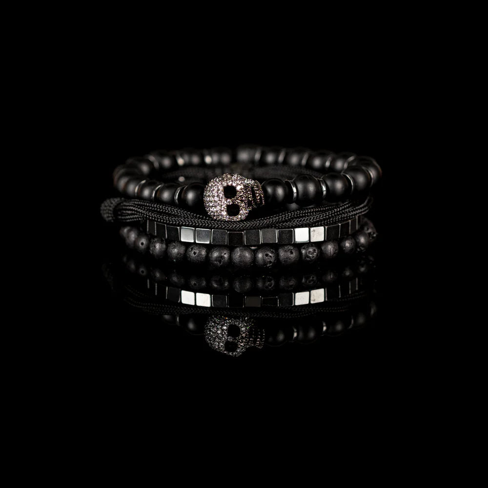 Men's handcrafted layered bracelet set featuring matte black onyx beads, black lava stone beads, polished silver hematite cube beads, black braided cord, and a crystal pavé skull centerpiece. Premium luxury men's stacked bracelet photographed on a clean white background.