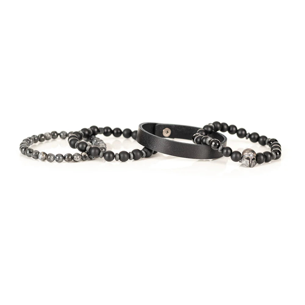 Men's handcrafted layered bracelet set featuring a black genuine leather cuff, matte black onyx beads, faceted black hematite beads, natural gray jasper gemstone beads, black hematite spacer beads, and a silver Spartan helmet centerpiece. Premium stacked men's bracelet photographed on a clean white background.
