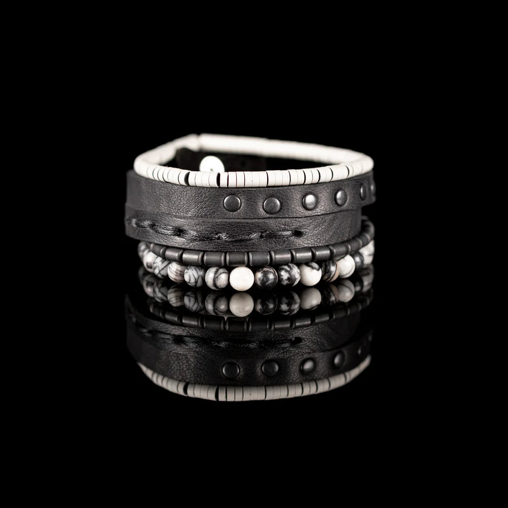 Men's handcrafted layered bracelet set featuring a black genuine leather wrap bracelet with gunmetal studs, white heishi disc beads, matte gunmetal hematite spacers, and natural black-and-white Picasso jasper gemstone beads. Premium stacked men's bracelet photographed on a clean white background.