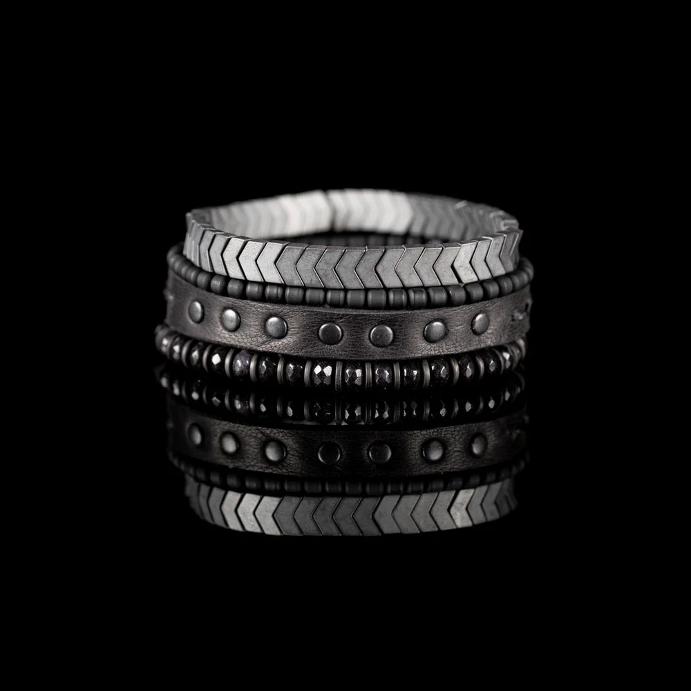 Men's handcrafted layered bracelet set featuring silver hematite arrow-shaped beads, black seed beads, a black leather wrap bracelet with matte gunmetal studs, and faceted black hematite beads. Premium minimalist men's bracelet stack photographed on a clean white background.
