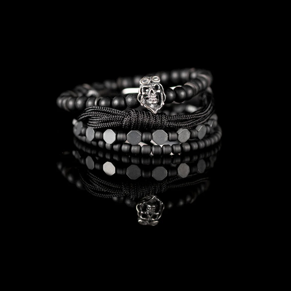 Men's handcrafted layered bracelet set featuring matte black onyx beads, black hematite hexagon beads, black seed beads, a black braided cord bracelet, and a silver aviator skull charm. Premium stacked men's bracelet photographed on a clean white background.