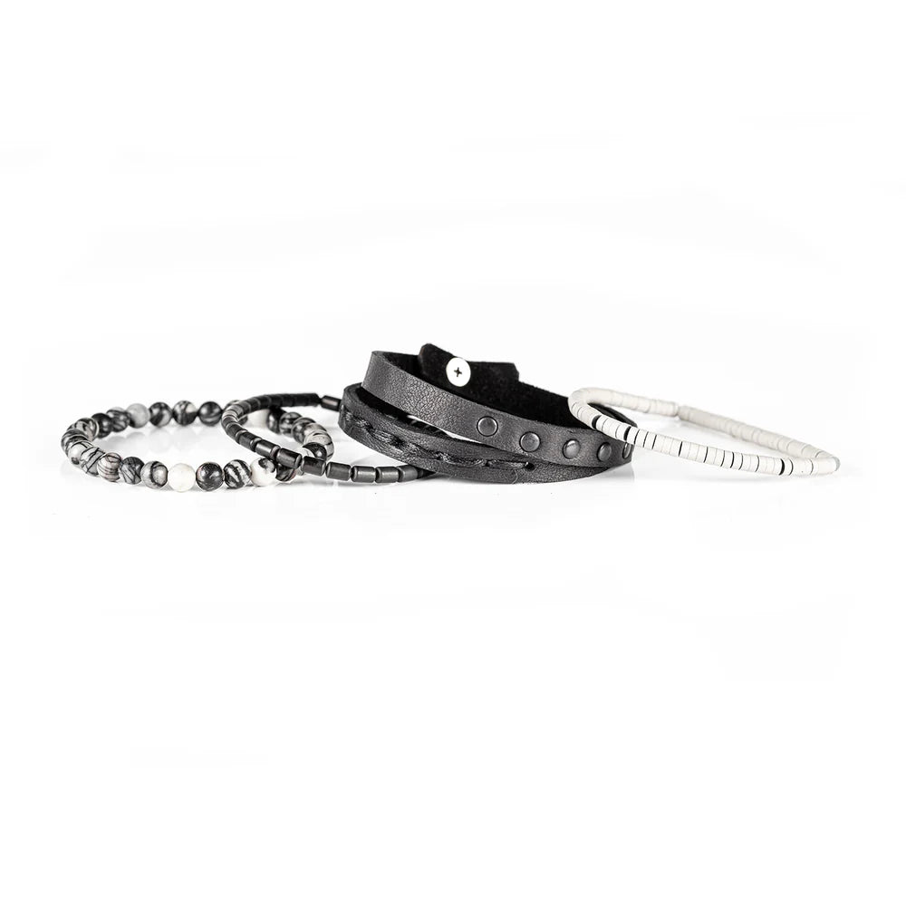 Men's handcrafted layered bracelet set featuring a black genuine leather wrap bracelet with gunmetal studs, white heishi disc beads, matte gunmetal hematite spacers, and natural black-and-white Picasso jasper gemstone beads. Premium stacked men's bracelet photographed on a clean white background.