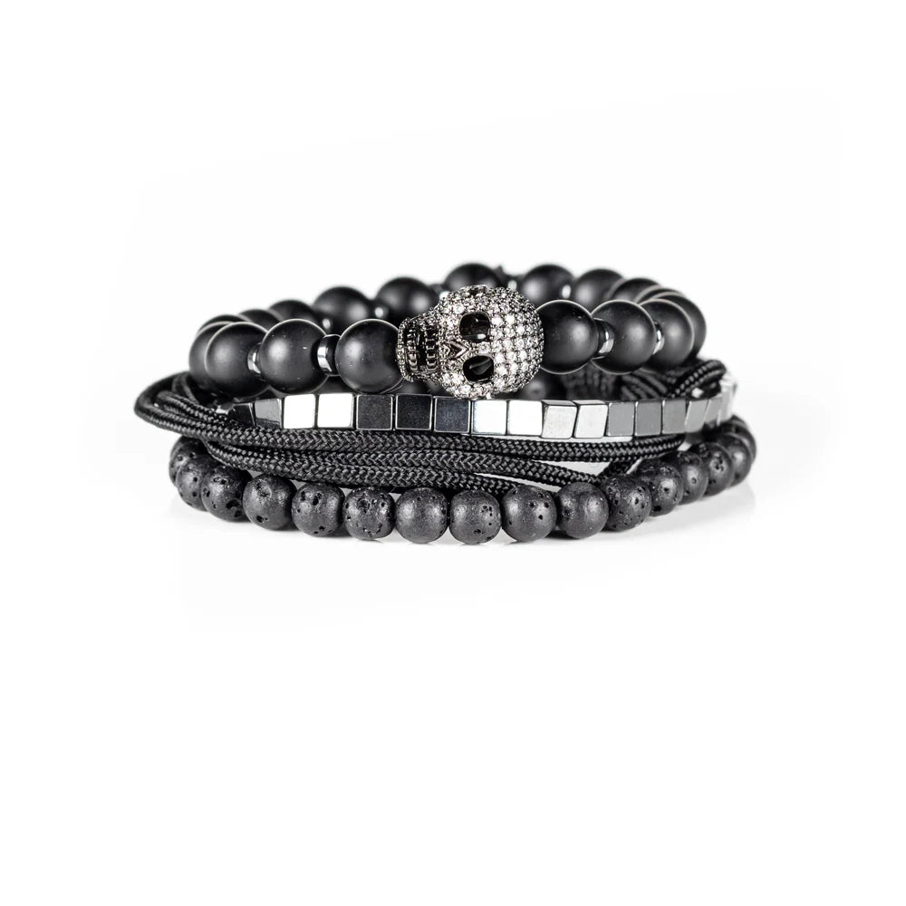 Men's handcrafted layered bracelet set featuring matte black onyx beads, black lava stone beads, polished silver hematite cube beads, black braided cord, and a crystal pavé skull centerpiece. Premium luxury men's stacked bracelet photographed on a clean white background.