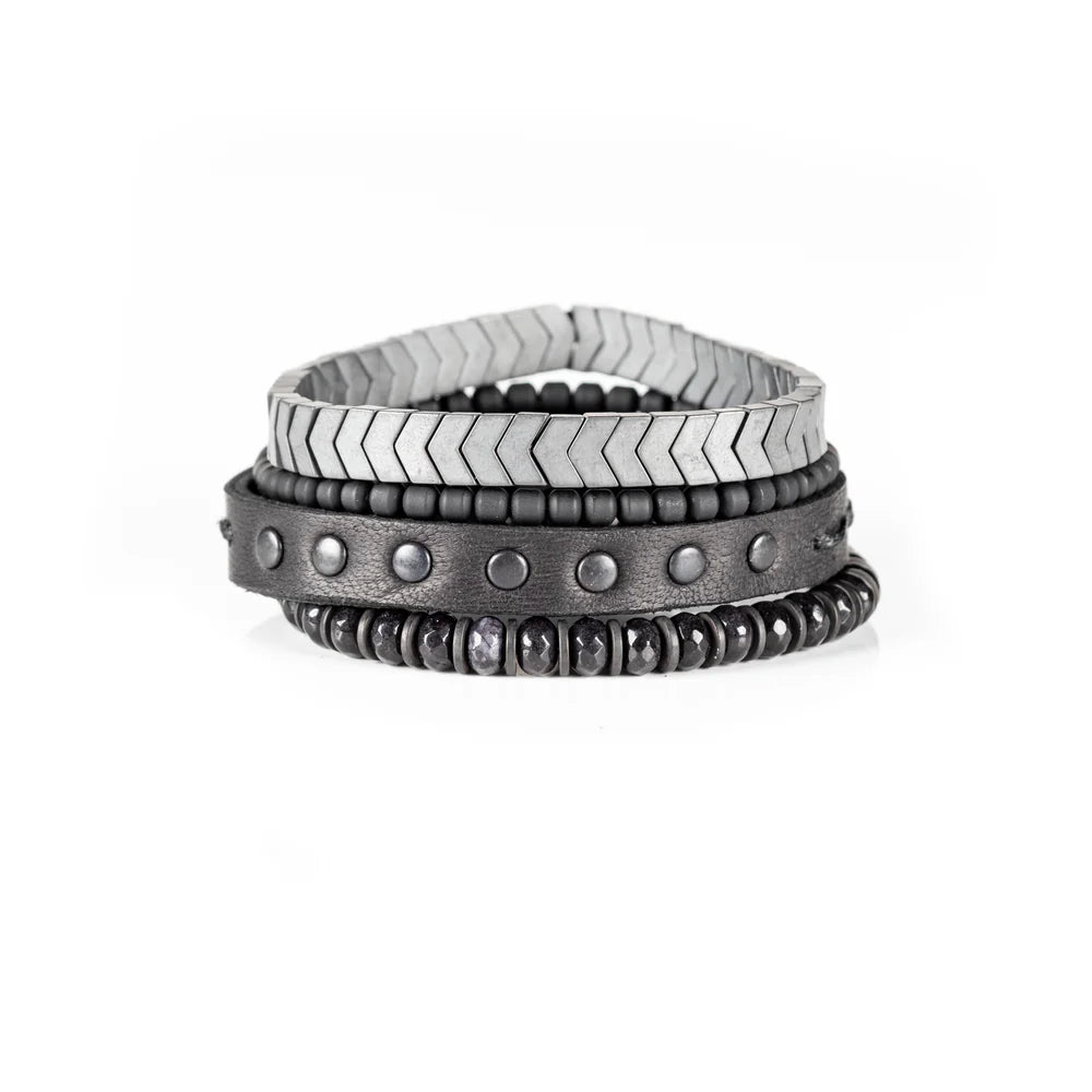 Men's handcrafted layered bracelet set featuring silver hematite arrow-shaped beads, black seed beads, a black leather wrap bracelet with matte gunmetal studs, and faceted black hematite beads. Premium minimalist men's bracelet stack photographed on a clean white background.