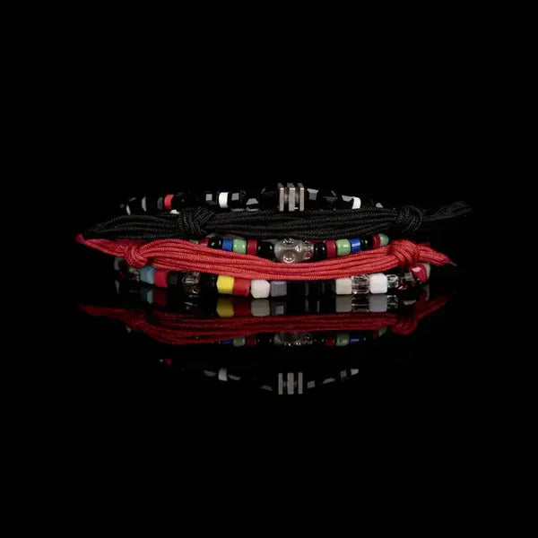 Layered men’s bracelet set featuring black and coral braided cord bracelets, multicolor bead accents, black faceted beads, and a silver-tone skull centerpiece, displayed on a white background.