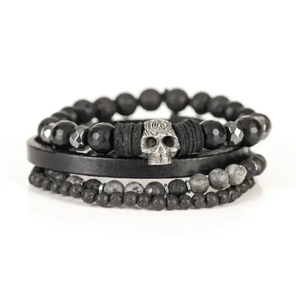 Layered men’s bracelet set featuring black faceted stone beads, a black leather band, lava stone beads, gray natural stone beads, and a silver skull centerpiece, displayed on a white background.