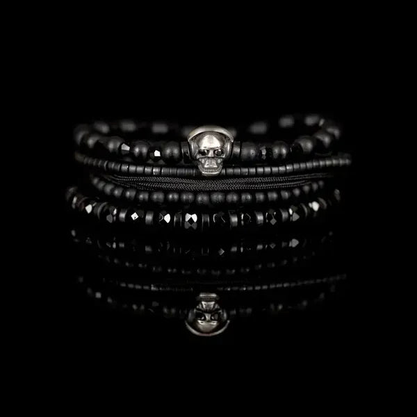 Black layered men’s bracelet with matte beads, faceted black stones, braided cord, and silver skull centerpiece on a white background.