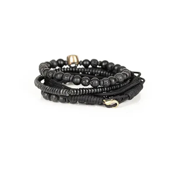 Black layered men’s bracelet with matte black beads, lava stone accents, braided cord wraps, and an antique silver skull centerpiece on a white background.