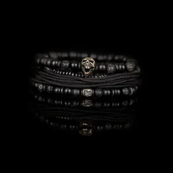 Black layered men’s bracelet with matte black beads, lava stone accents, braided cord wraps, and an antique silver skull centerpiece on a white background.