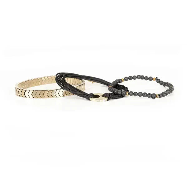 Black layered men’s bracelet featuring lava stone beads, braided cord wraps, a gold-tone centerpiece ring, and gold chevron metal accents on a white background.