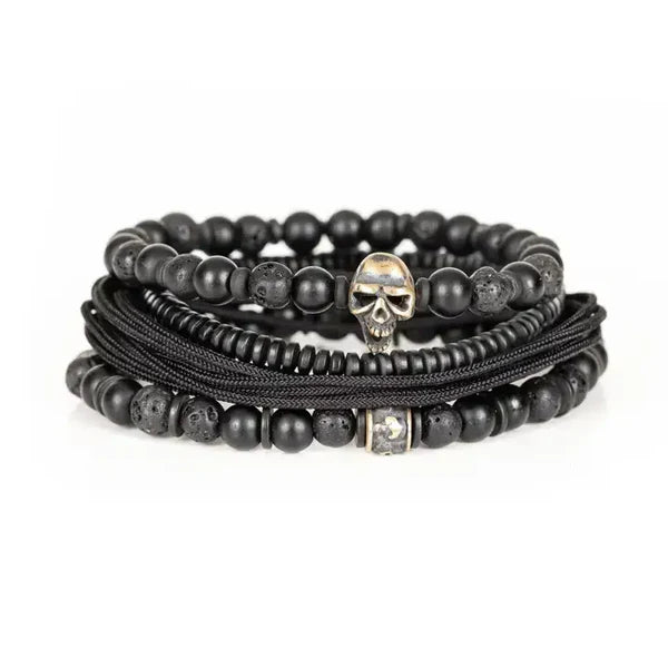 Black layered men’s bracelet with matte black beads, lava stone accents, braided cord wraps, and an antique silver skull centerpiece on a white background.