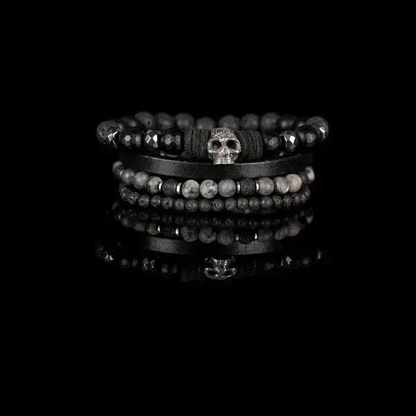 Layered men’s bracelet set featuring black faceted stone beads, a black leather band, lava stone beads, gray natural stone beads, and a silver skull centerpiece, displayed on a white background.