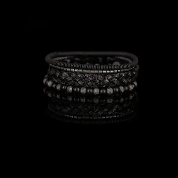 Layered men’s bracelet set featuring a braided black leather band, matte black beads, faceted hematite beads, and hematite disc accents in a monochromatic stacked design, displayed on a white background.