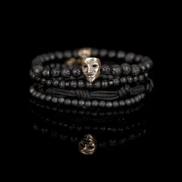 Black layered men’s bracelet featuring lava stone beads, faceted black stone accents, braided cord wraps, and an antique gold theatrical mask centerpiece on a white background.