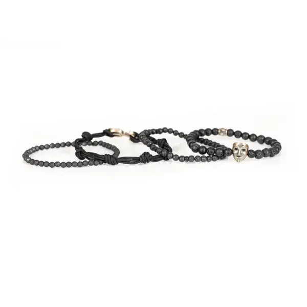 Black layered men’s bracelet featuring lava stone beads, faceted black stone accents, braided cord wraps, and an antique gold theatrical mask centerpiece on a white background.