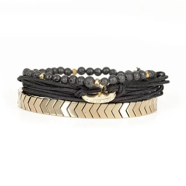 Black layered men’s bracelet featuring lava stone beads, braided cord wraps, a gold-tone centerpiece ring, and gold chevron metal accents on a white background.