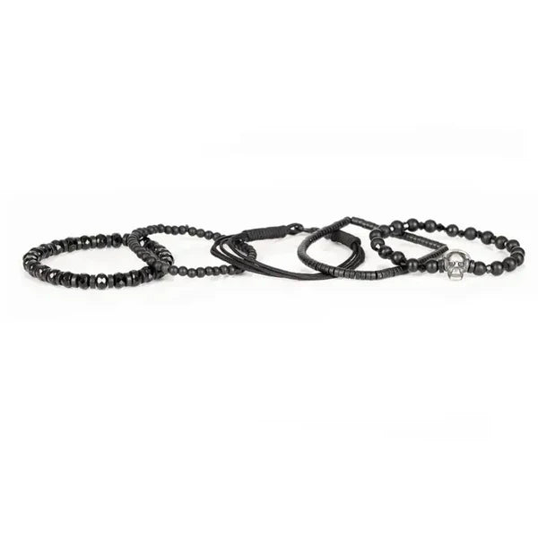 Black layered men’s bracelet with matte beads, faceted black stones, braided cord, and silver skull centerpiece on a white background.
