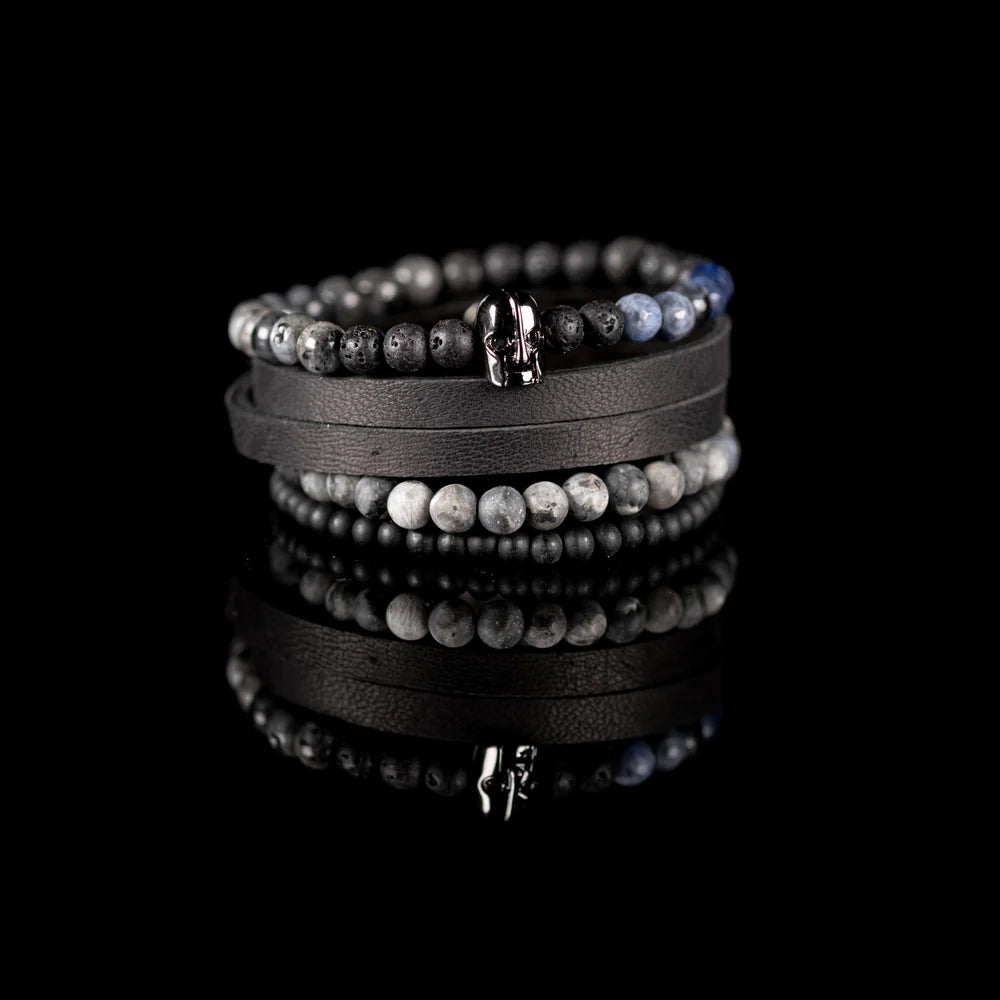 Men's handcrafted black leather wrap bracelet set featuring matte black onyx beads, natural gray jasper stone beads, black lava stone, blue sodalite accent beads, and a polished black skull centerpiece. Premium layered men's bracelet stack photographed on a clean white background.