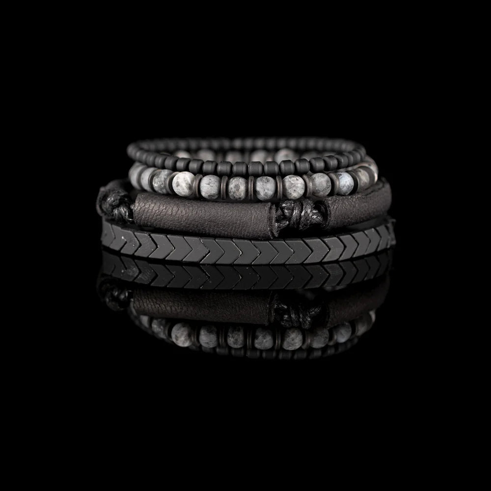 Men's handcrafted layered bracelet set featuring black leather wrap bands, matte black hematite arrow beads, black seed beads, and natural gray jasper gemstone beads. Premium stacked men's bracelet with minimalist design photographed on a clean white background.