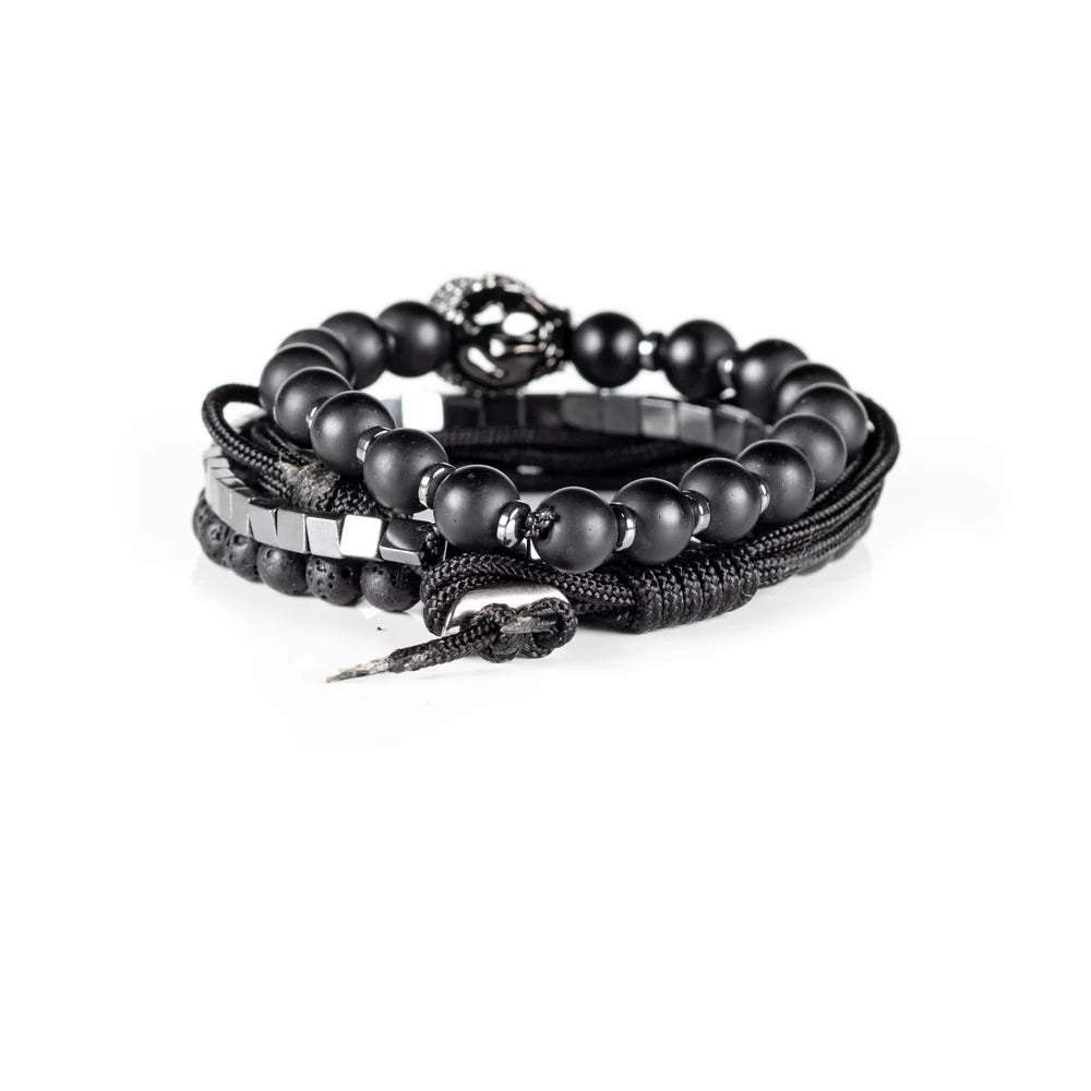 Men's handcrafted layered bracelet set featuring matte black onyx beads, black lava stone beads, polished silver hematite cube beads, black braided cord, and a crystal pavé skull centerpiece. Premium luxury men's stacked bracelet photographed on a clean white background.