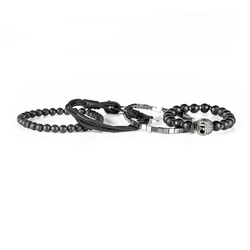 Men's handcrafted layered bracelet set featuring matte black onyx beads, black lava stone beads, polished silver hematite cube beads, black braided cord, and a crystal pavé skull centerpiece. Premium luxury men's stacked bracelet photographed on a clean white background.