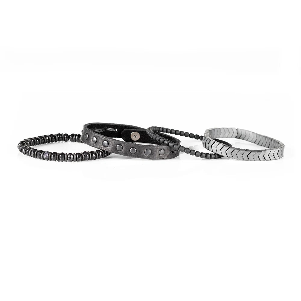 Men's handcrafted layered bracelet set featuring silver hematite arrow-shaped beads, black seed beads, a black leather wrap bracelet with matte gunmetal studs, and faceted black hematite beads. Premium minimalist men's bracelet stack photographed on a clean white background.