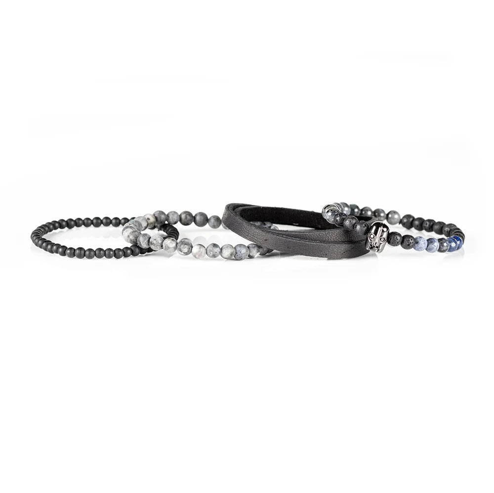 Men's handcrafted black leather wrap bracelet set featuring matte black onyx beads, natural gray jasper stone beads, black lava stone, blue sodalite accent beads, and a polished black skull centerpiece. Premium layered men's bracelet stack photographed on a clean white background.