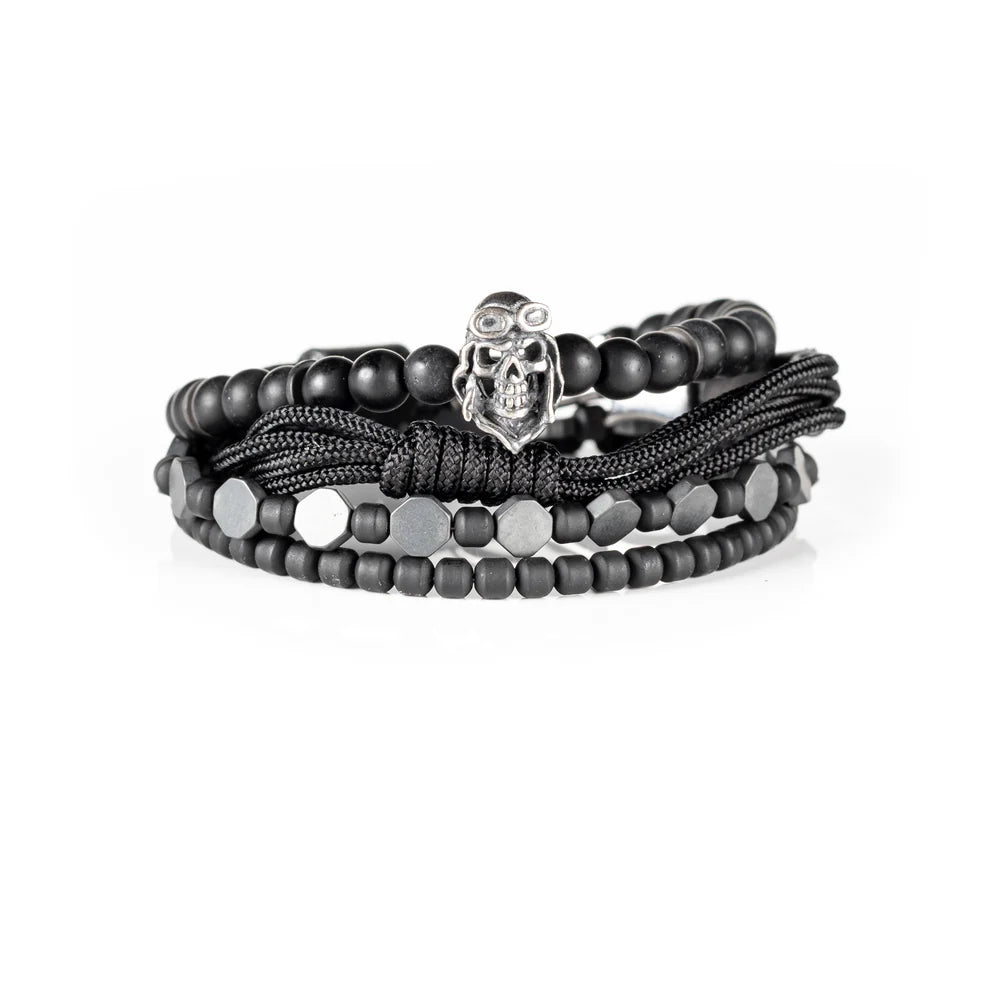 Men's handcrafted layered bracelet set featuring matte black onyx beads, black hematite hexagon beads, black seed beads, a black braided cord bracelet, and a silver aviator skull charm. Premium stacked men's bracelet photographed on a clean white background.