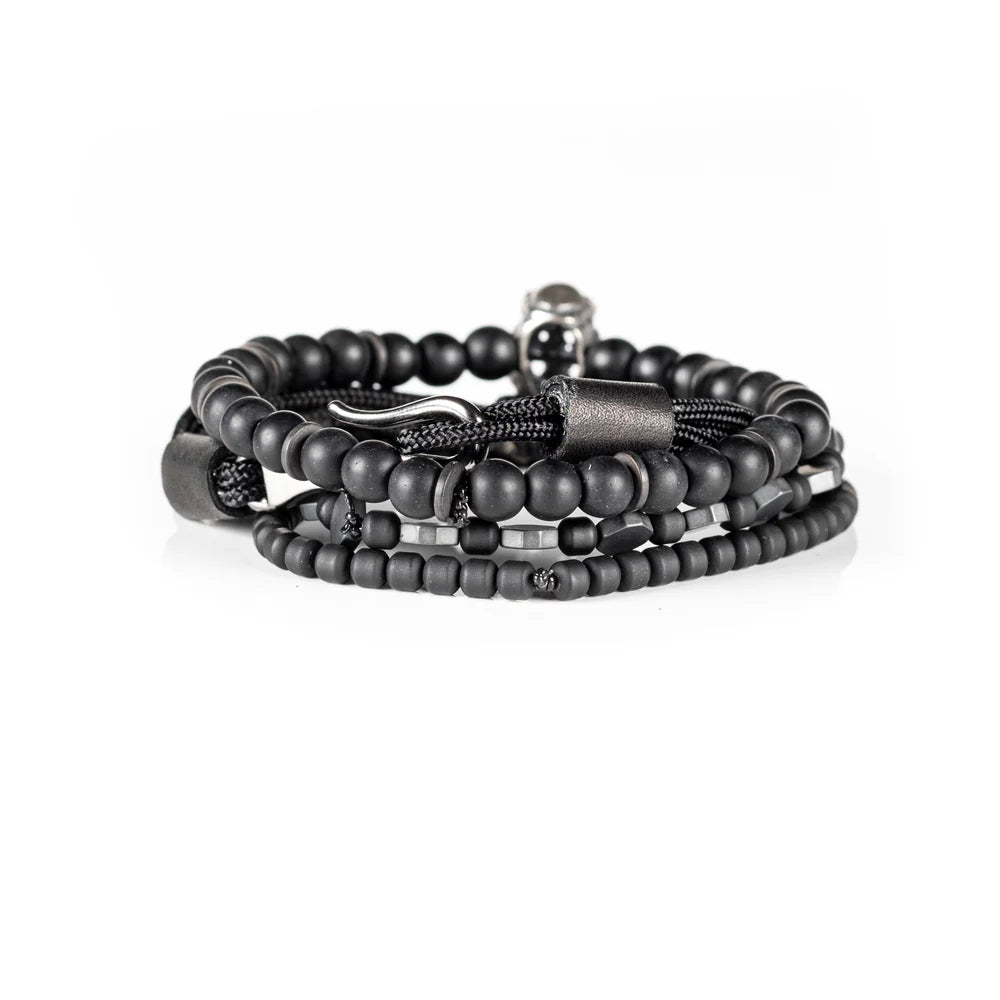 Men's handcrafted layered bracelet set featuring matte black onyx beads, black hematite hexagon beads, black seed beads, a black braided cord bracelet, and a silver aviator skull charm. Premium stacked men's bracelet photographed on a clean white background.