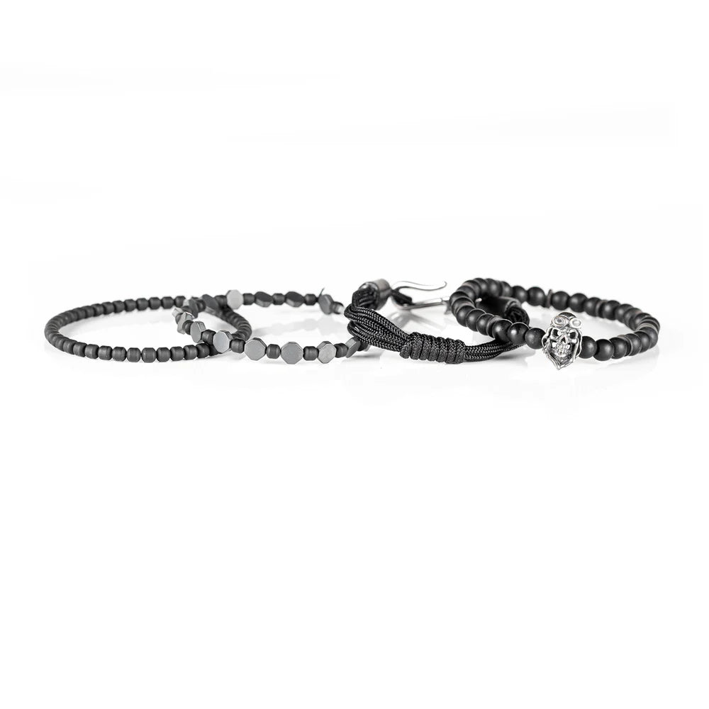 Men's handcrafted layered bracelet set featuring matte black onyx beads, black hematite hexagon beads, black seed beads, a black braided cord bracelet, and a silver aviator skull charm. Premium stacked men's bracelet photographed on a clean white background.
