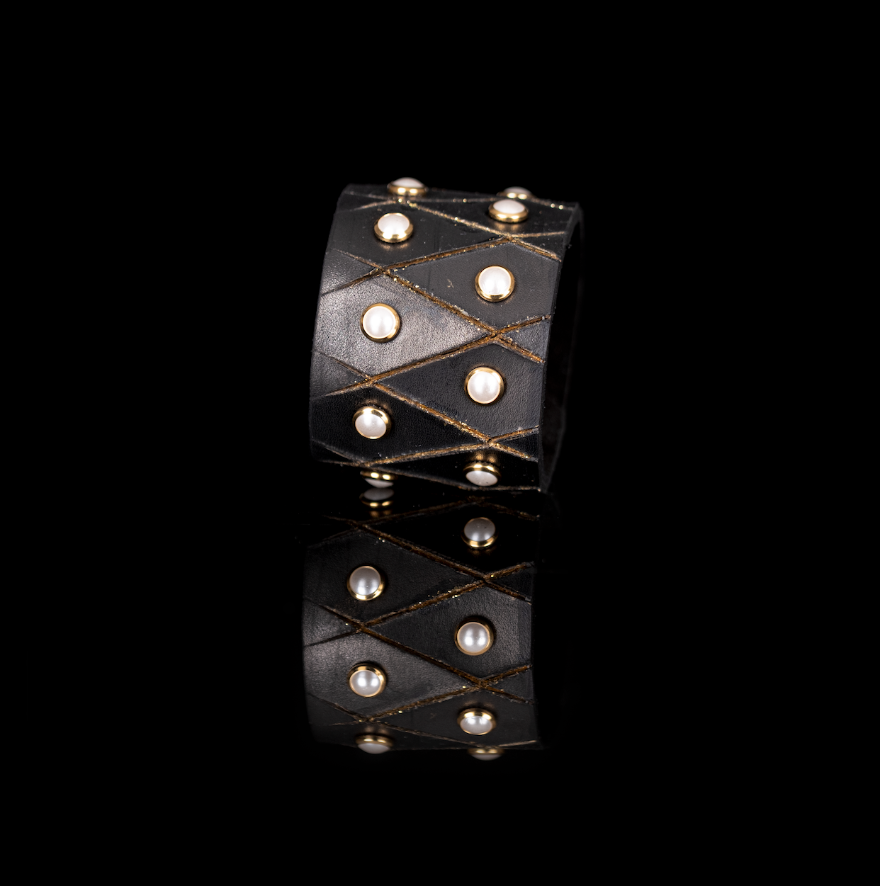 Black leather cuff bracelet with geometric stitched pattern and gold-tone studs featuring pearl accents, wide statement women’s bracelet on dark background